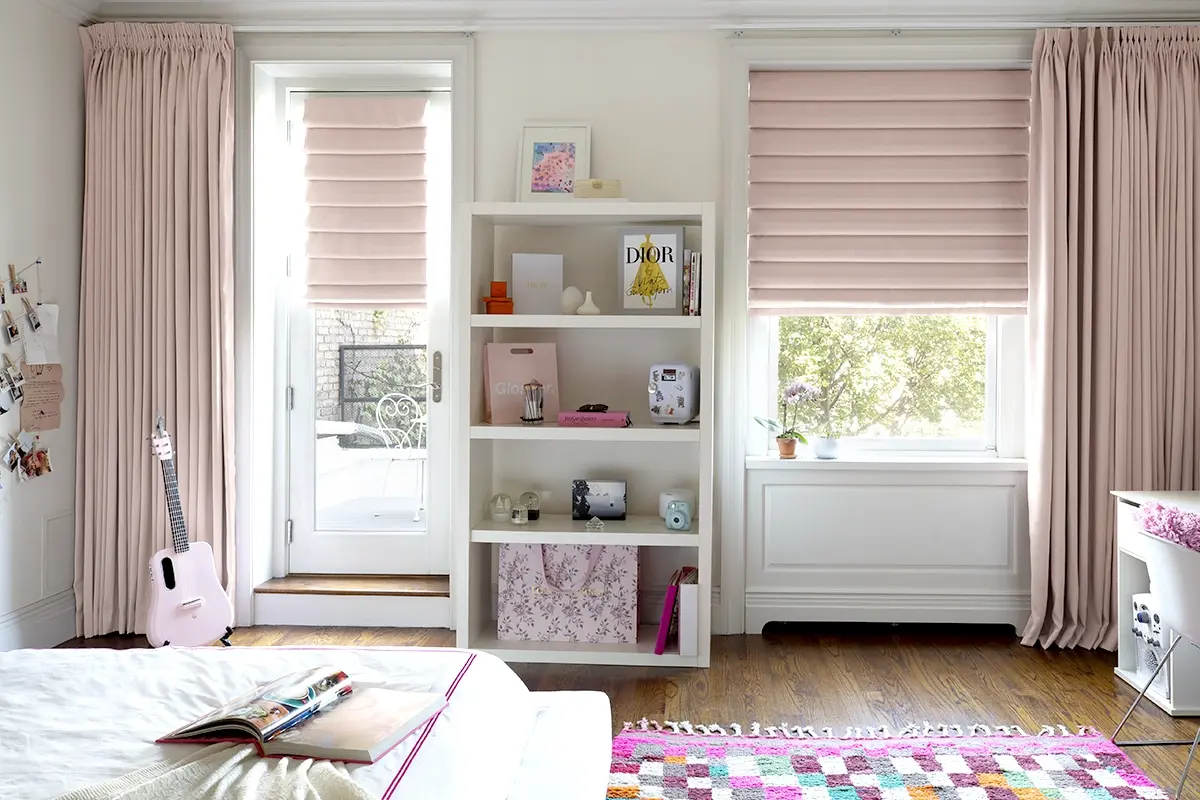A child's room features pink roman shades and pink drapery layered on two adjacent windows.