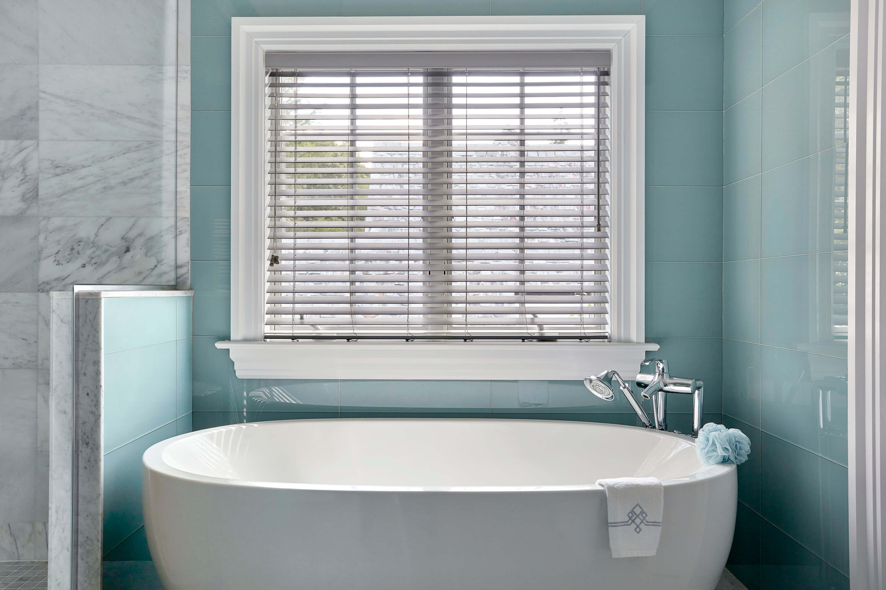 The modern bathroom features a large soaking tub, light grey cordless faux wood blinds, and blue and white tiles.