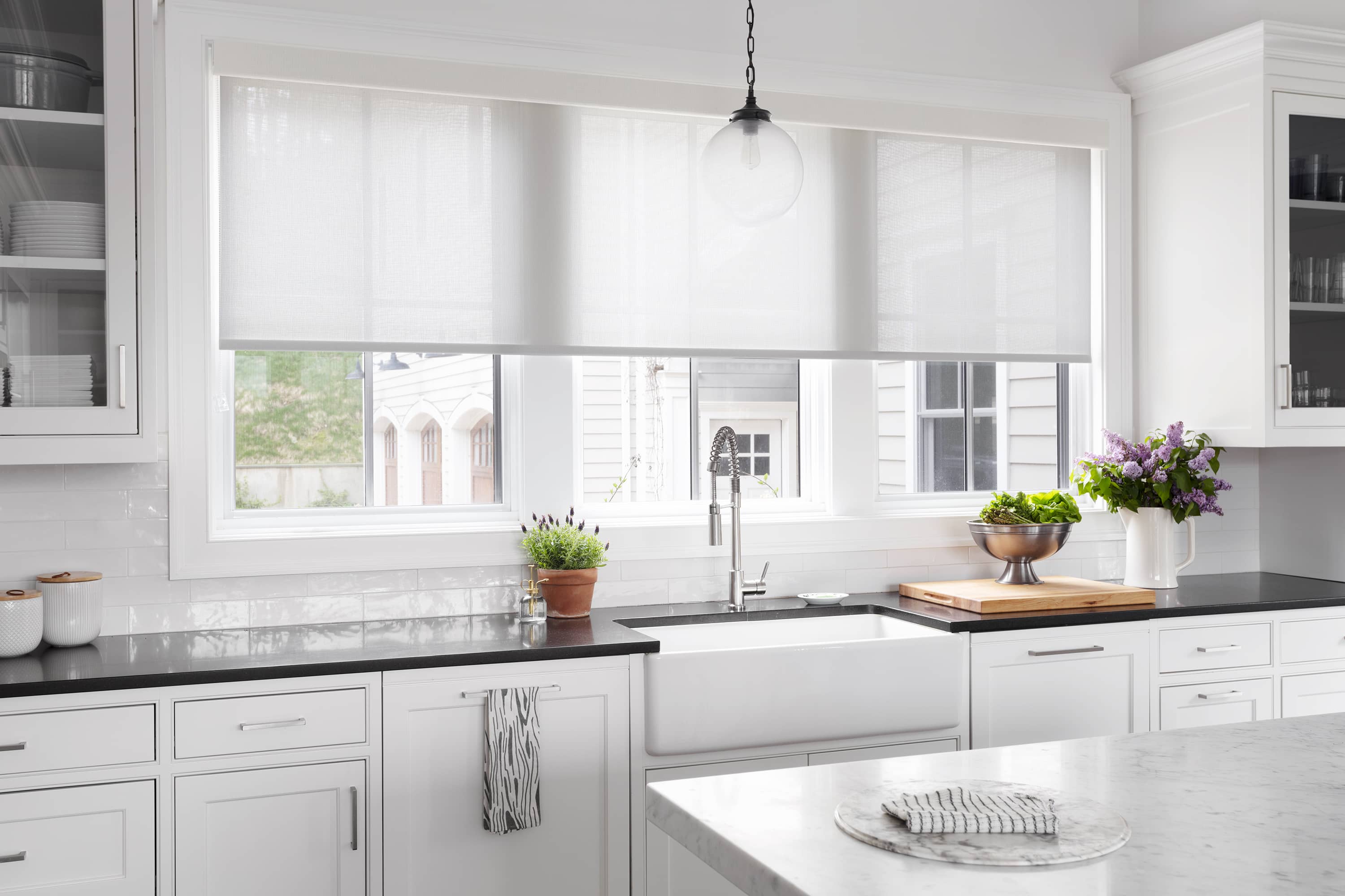 A white, cordless roller shades with 10% openness covers three large windows above a kitchen sink.