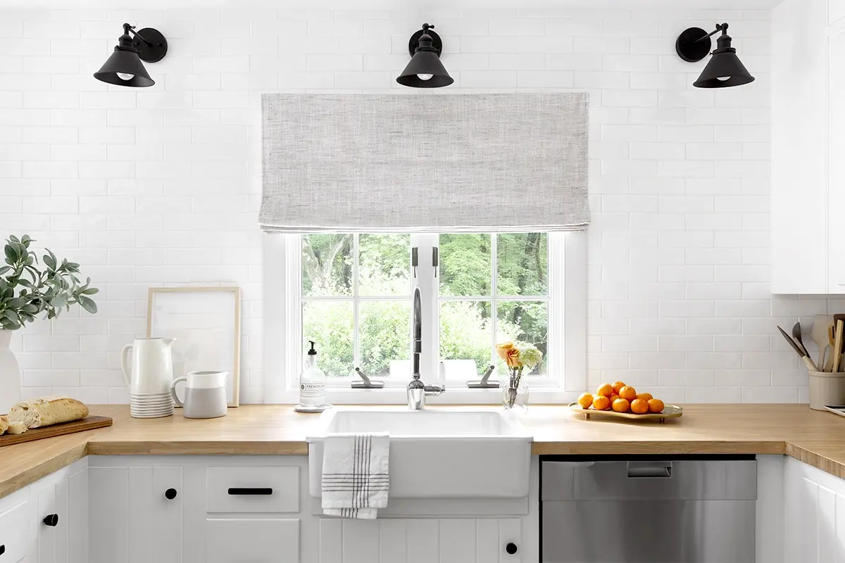 A white Roman shade with subtle flecks of grey and flat style pleat adorns a large window of a sink in a modern kitchen.