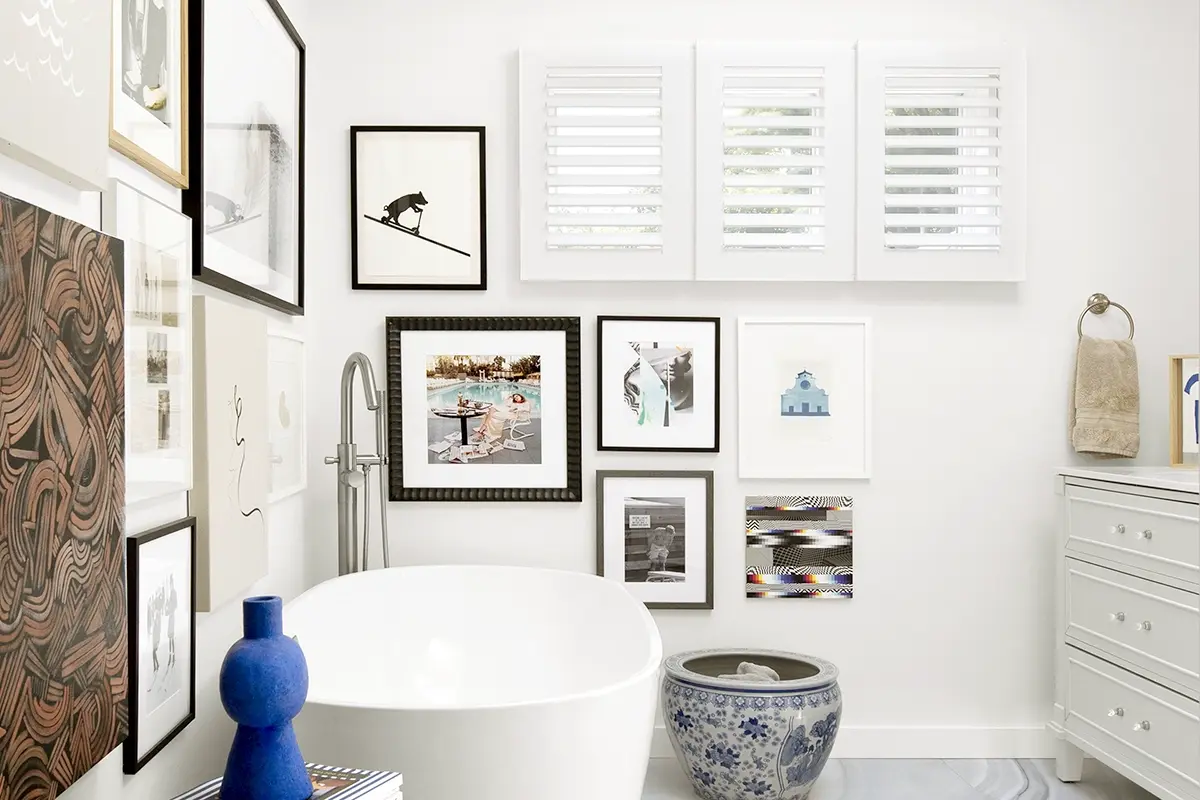 A modern and eclectic bathroom with a freestanding tub and a gallery of many artworks on the wall features shutters on a high window.