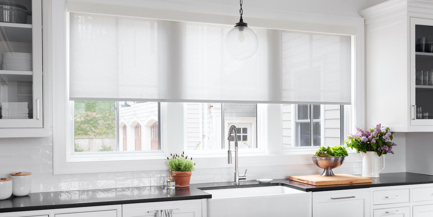 White roller shades cover a large window over a kitchen sink.