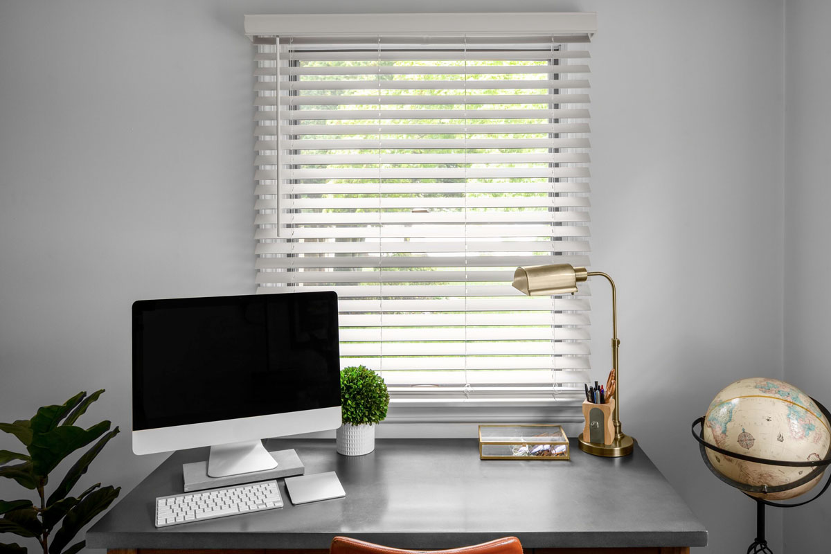 Classic white faux wood blinds cover a small window in a home office.
