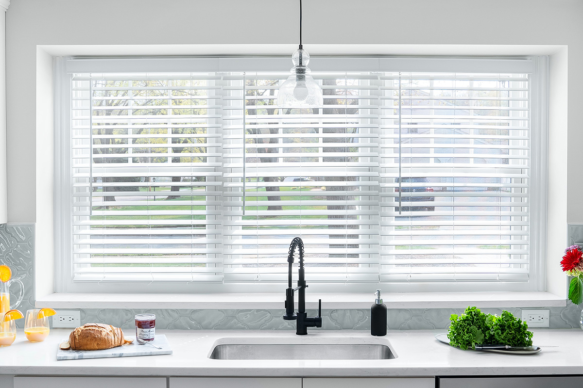 Three side-by-side windows above a kitchen sink feature white faux wood blinds.