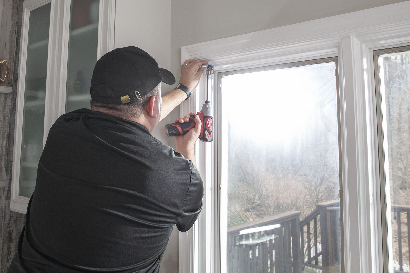 A man in a black hat and black polo shirt uses a drill to screw a roman shade bracket into the top of a kitchen window frame.
