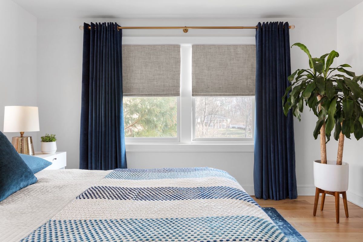 Blue velvet drapes are paired with tan linen roman shades on two large windows in a modern bedroom.