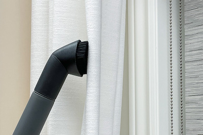 A vacuum attachment with bristles being used to clean drapes.