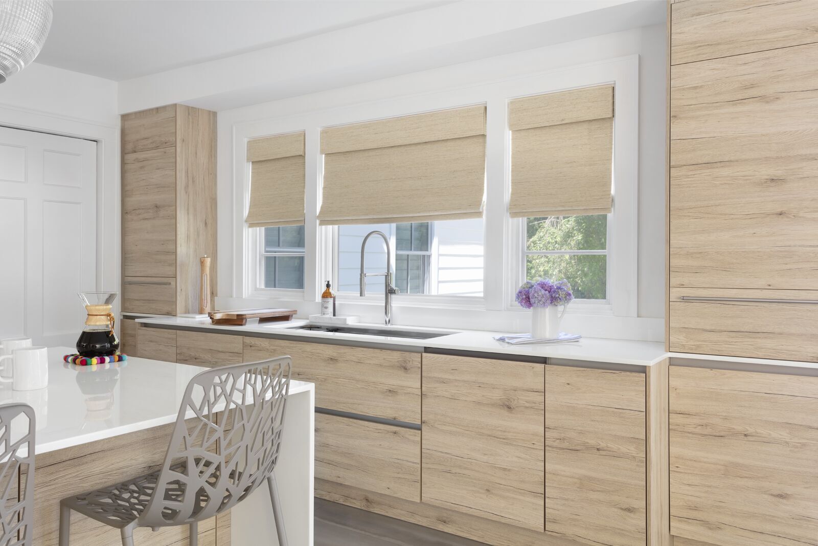Tan woven shades cover three windows in a modern kitchen with cabinets that match the same shade as the shades.