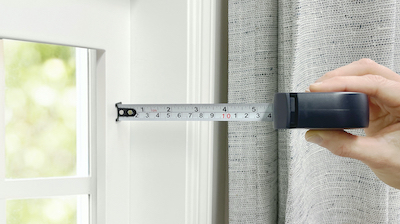 A hand holds a measuring tape against a window's right side jamb.
