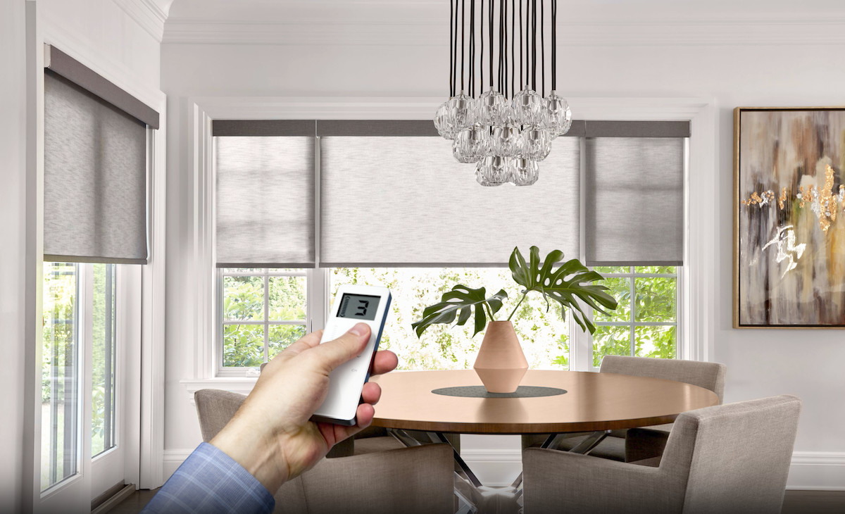 A hand points a remote at a modern dining room with large windows featuring roller shades.