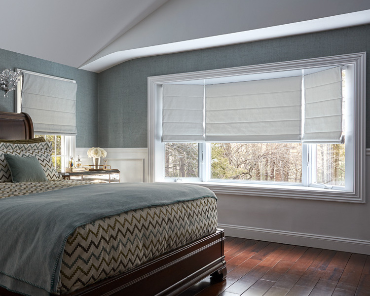A bay window in a spacious bedroom is covered with white roman shades.