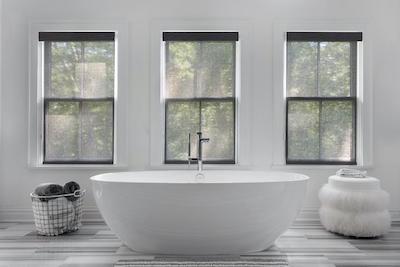 A large contemporary bathtub is situated under three large windows featuring solar shades.