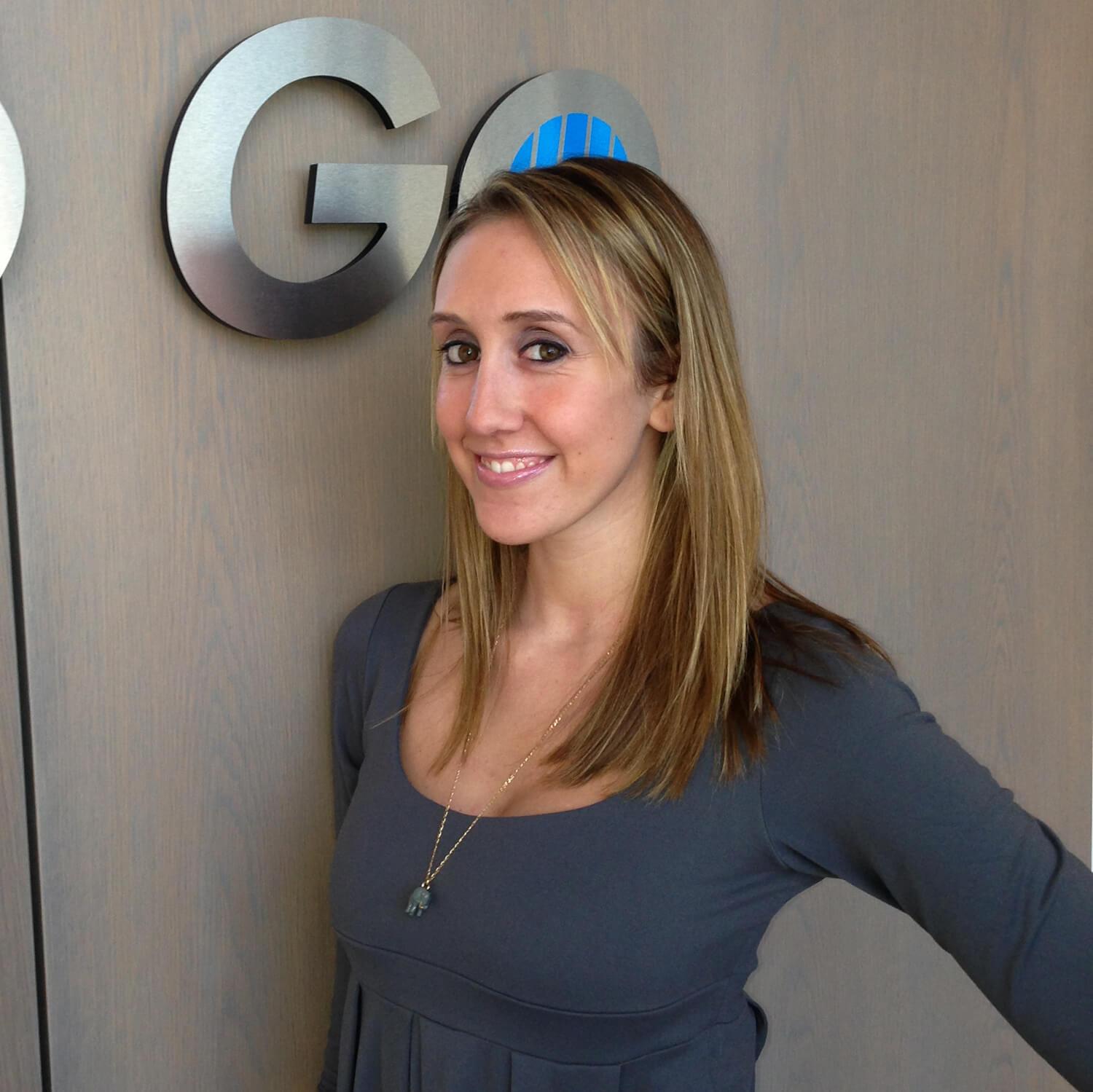 A woman smiles in front of a large Blinds To Go sign at the BTG headquarters