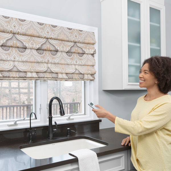 A woman uses a remote control to raise a classic pleat, roman shade in a modern kitchen.