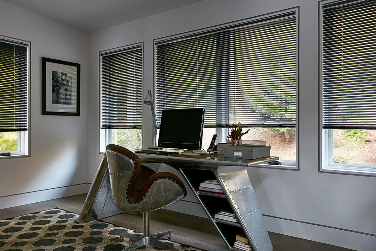 Dark blue aluminum blinds cover a large window over a large desk in a home office.