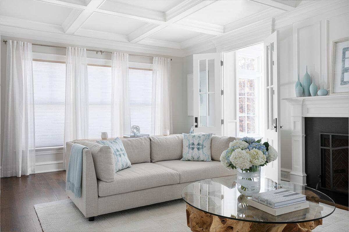 A living room with light grey textured sheer drapes and antique white cellular shades