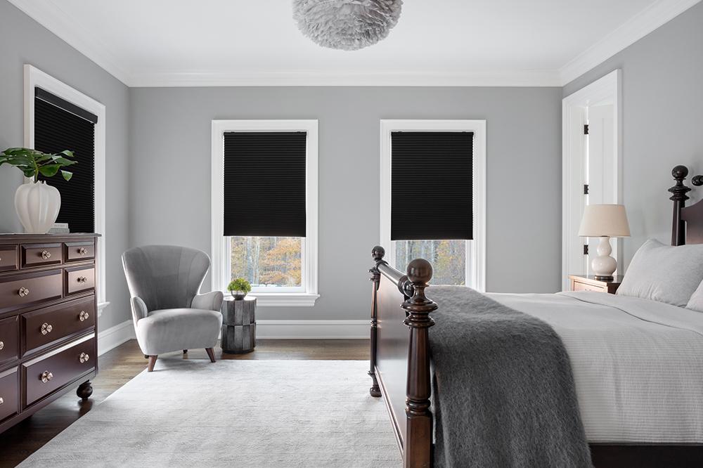 A contemporary bedroom in light grey tones has three windows partially covered by black room-darkening roller shades.