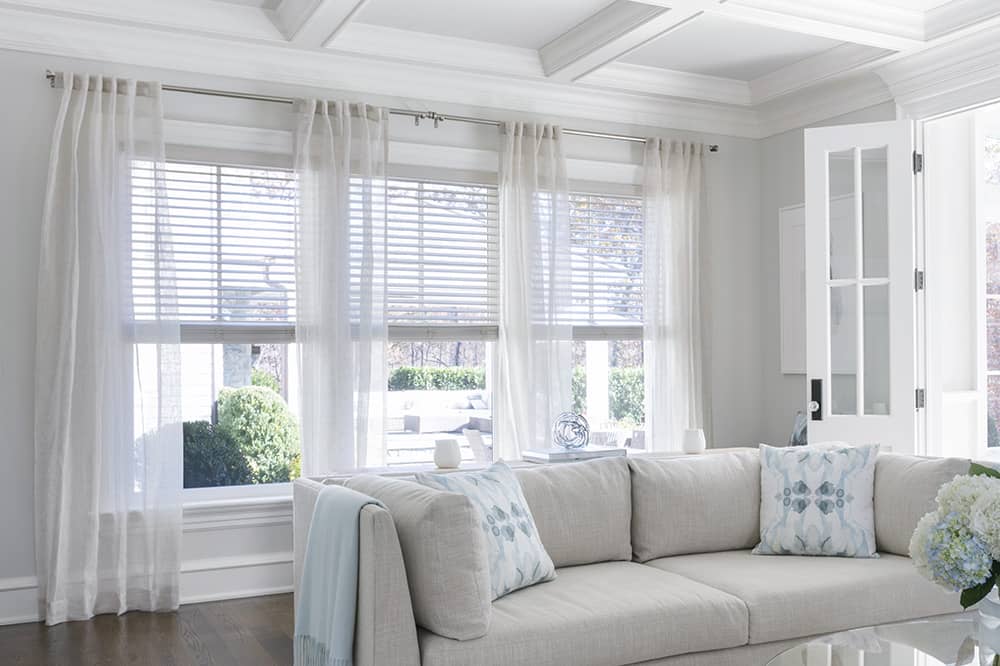 Sheer curtains cover three windows in a bright, spacious living room.