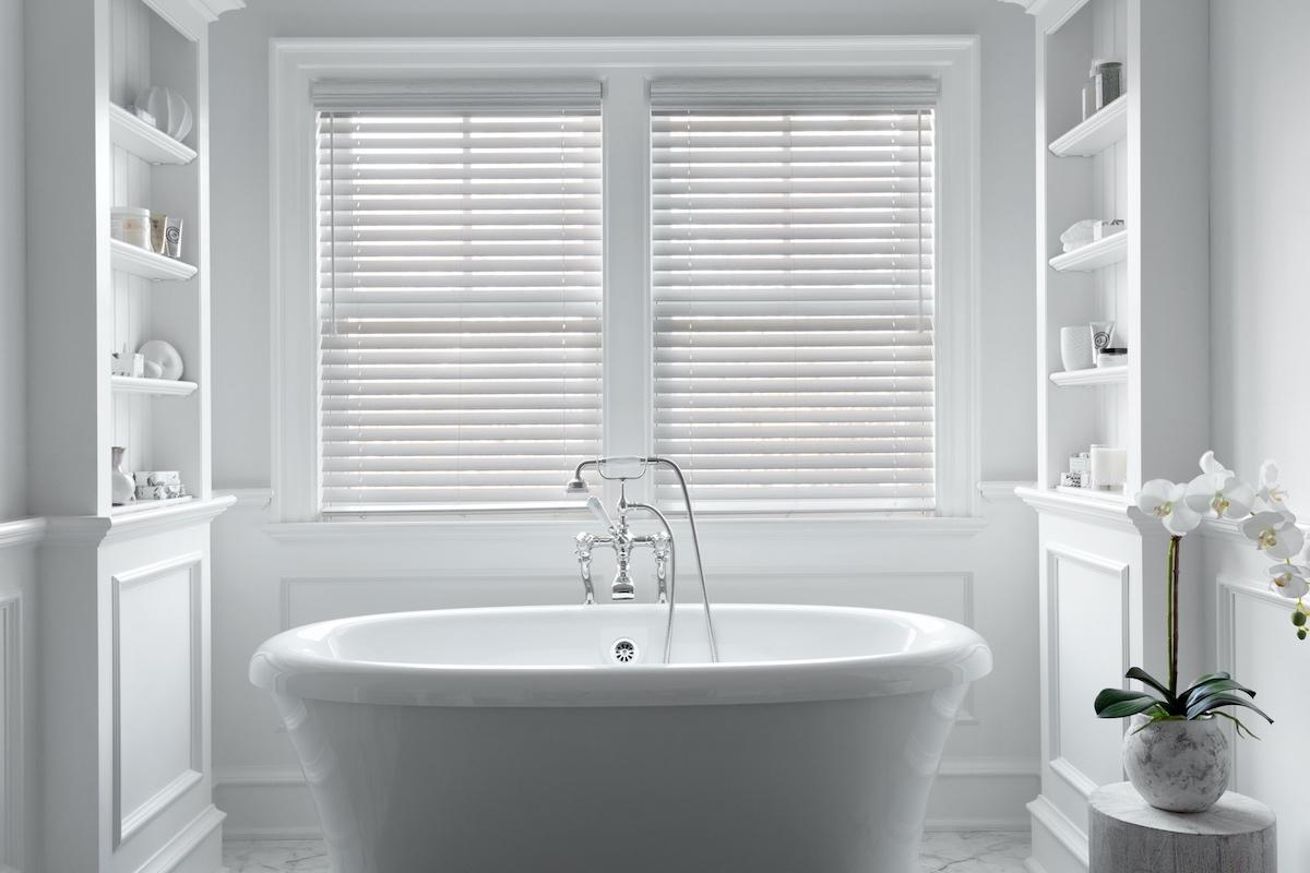 Stores en similibois blancs sur deux fenêtres à l’arrière d’une grande baignoire dans une salle de bain moderne.