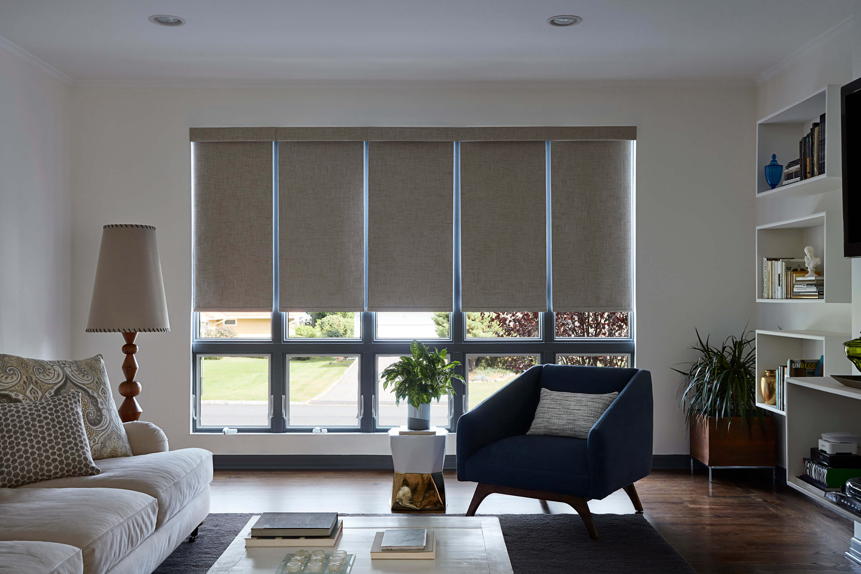 Five motorized tan blackout roller shades on side-by-side windows are synched in height in a modern living room.