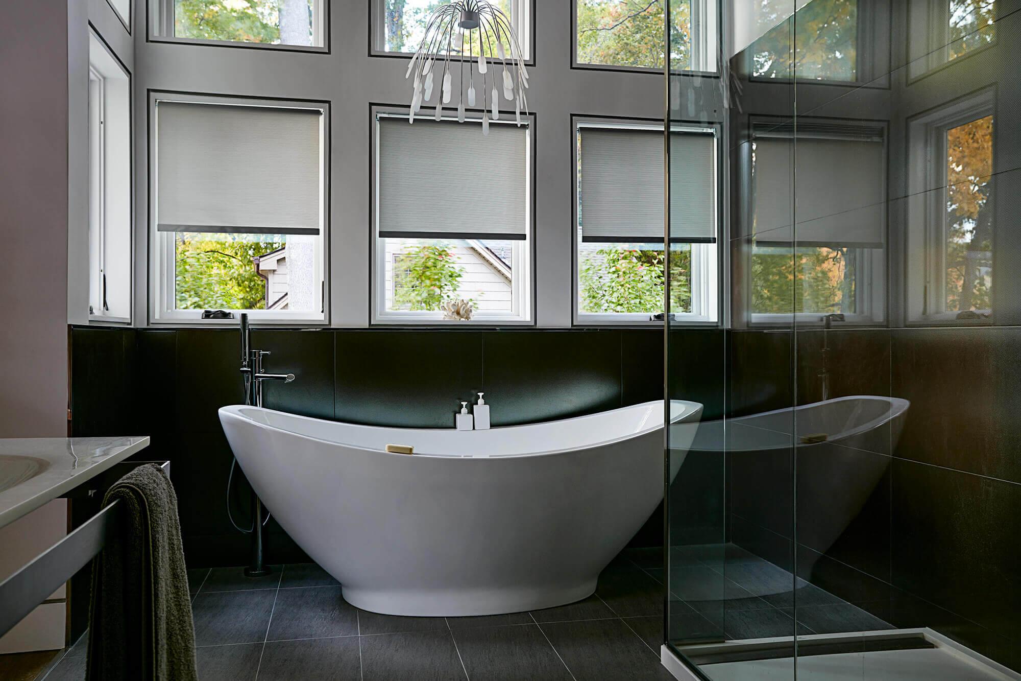 A modern bathroom with a free standing tub and glass shower has three windows covered with roller shades behind the tub