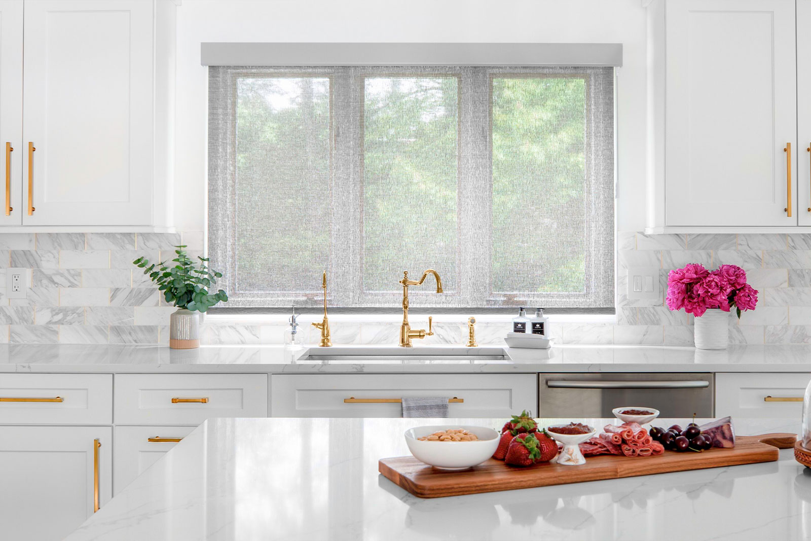 Light shines through light grey solar shade on three windows behind a kitchen sink.