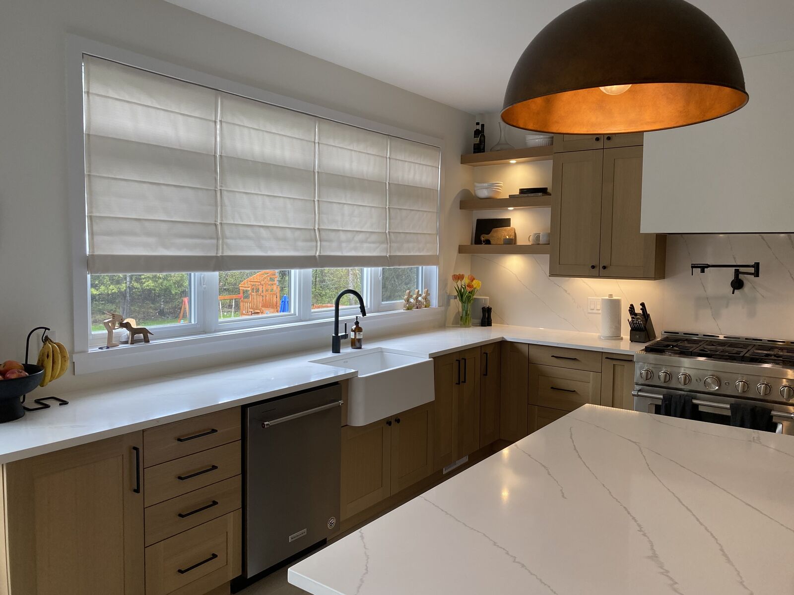 A modern kitchen features four windows partially covered with flat roman shades.