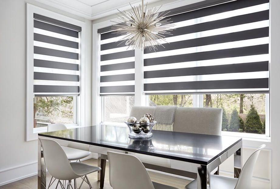 A modern dining room with large windows covered with dark grey and transparent striped zebra shades.
