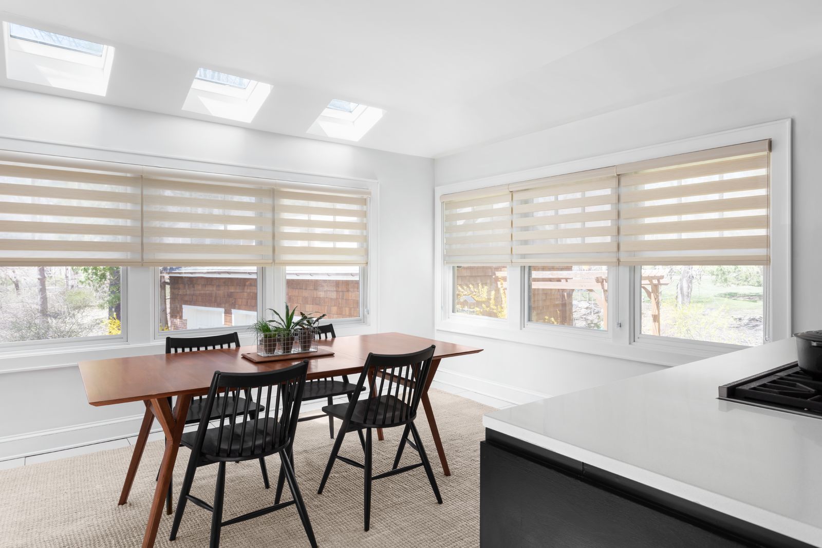 A modern dining area and kitchen with large windows covered in light tan Cascade sheer shades