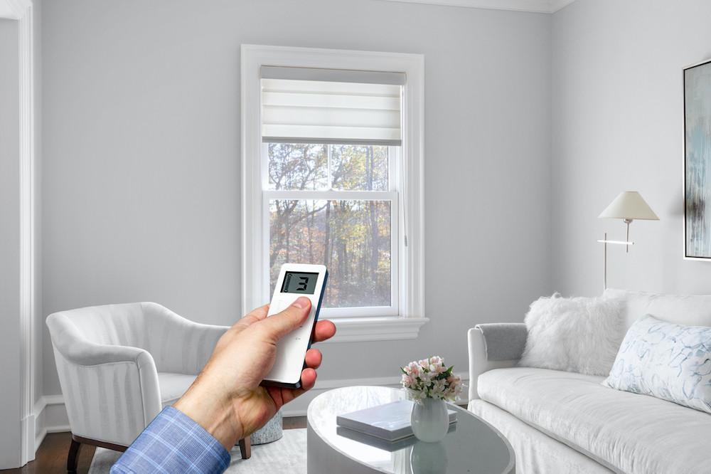 A window with a shade mostly open in a beautiful contemporary living room, with a hand holding a remote in the foreground.