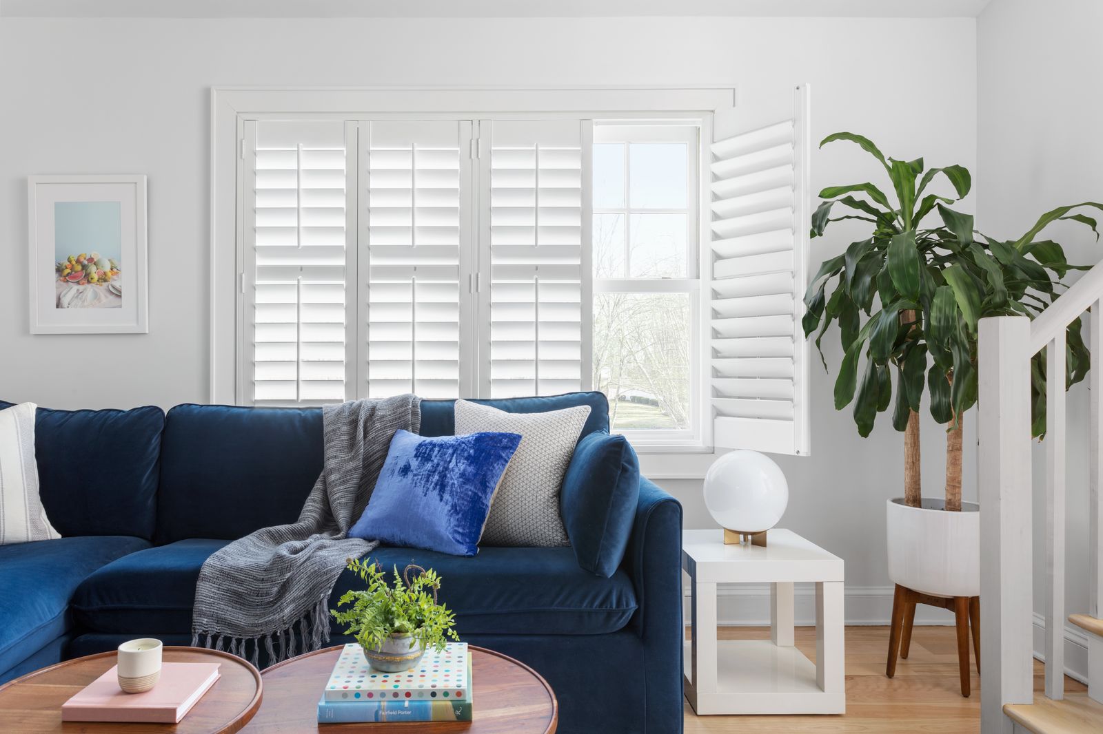 A trendy living room with a blue couch glowing by sunlight streaming through white Plantation shutters.
