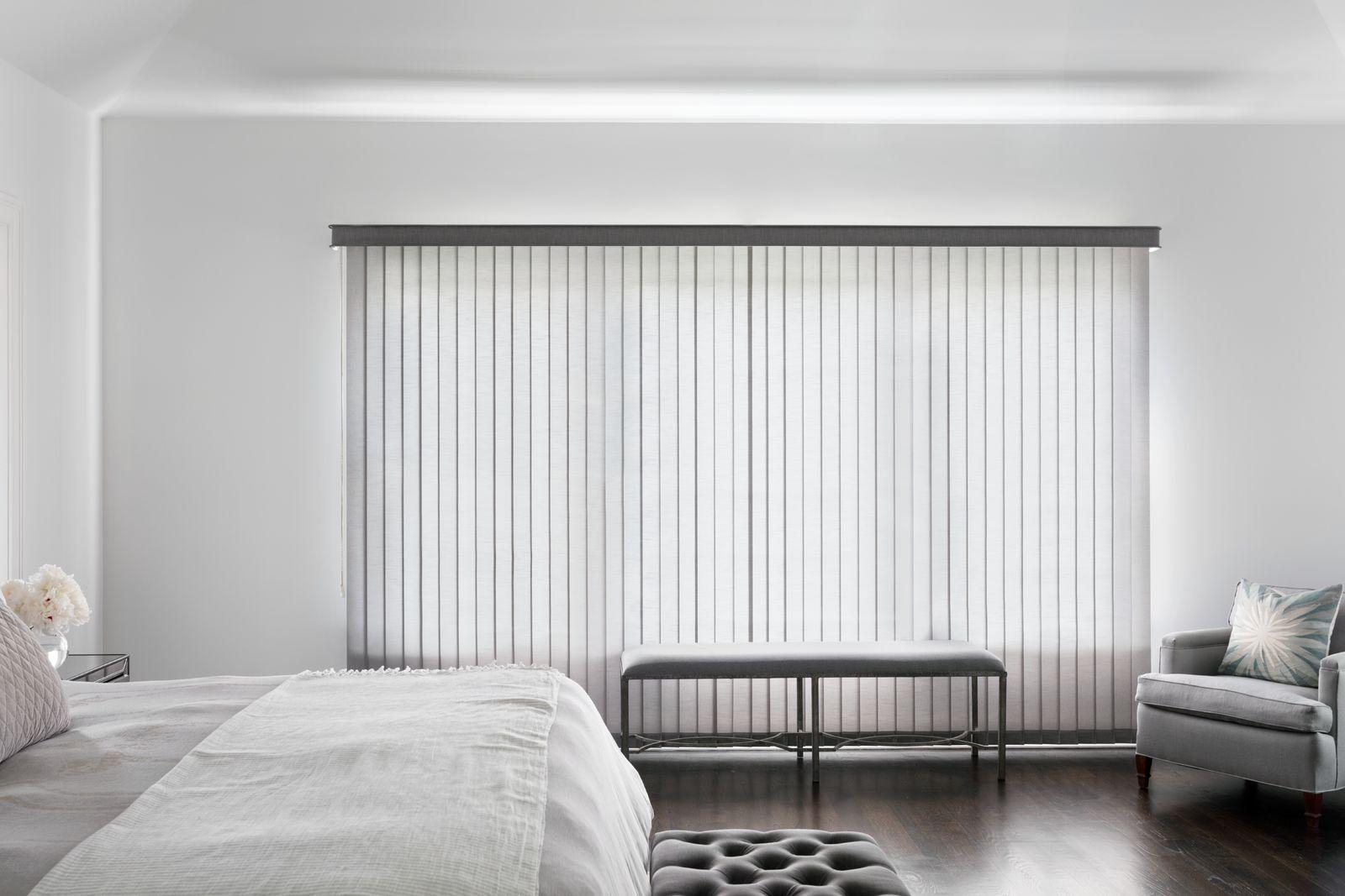 Des stores verticaux en tissu gris avec des lattes fermées de façon à permettre à une lumière diffuse de baigner une grande chambre à coucher de style contemporain.