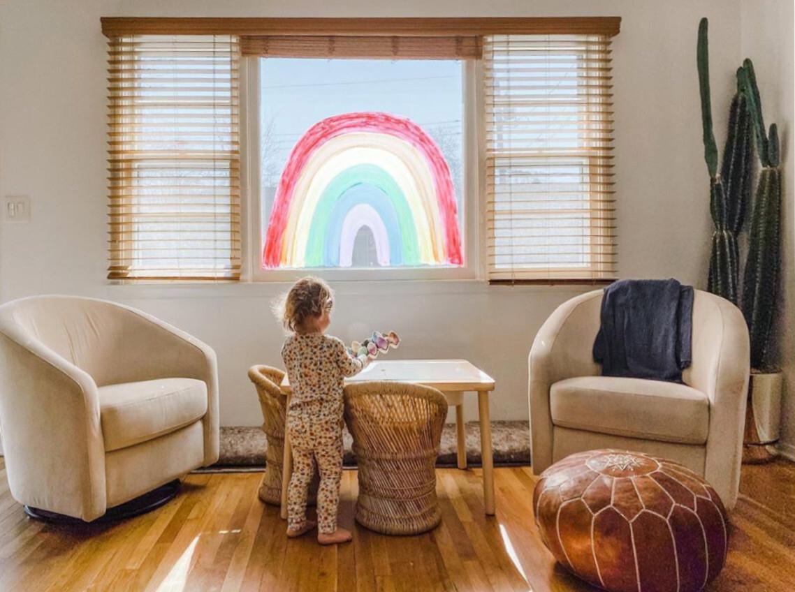 A bay window features cordless wood blinds with the middle blind in the up position revealing a rainbow painted on the window, while a toddler plays in the foreground.