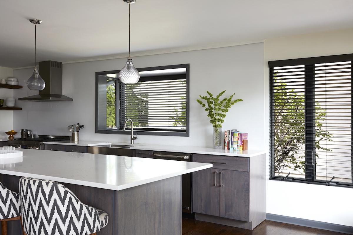 A modern kitchen in white and grey tones features two windows with black real wood blinds.