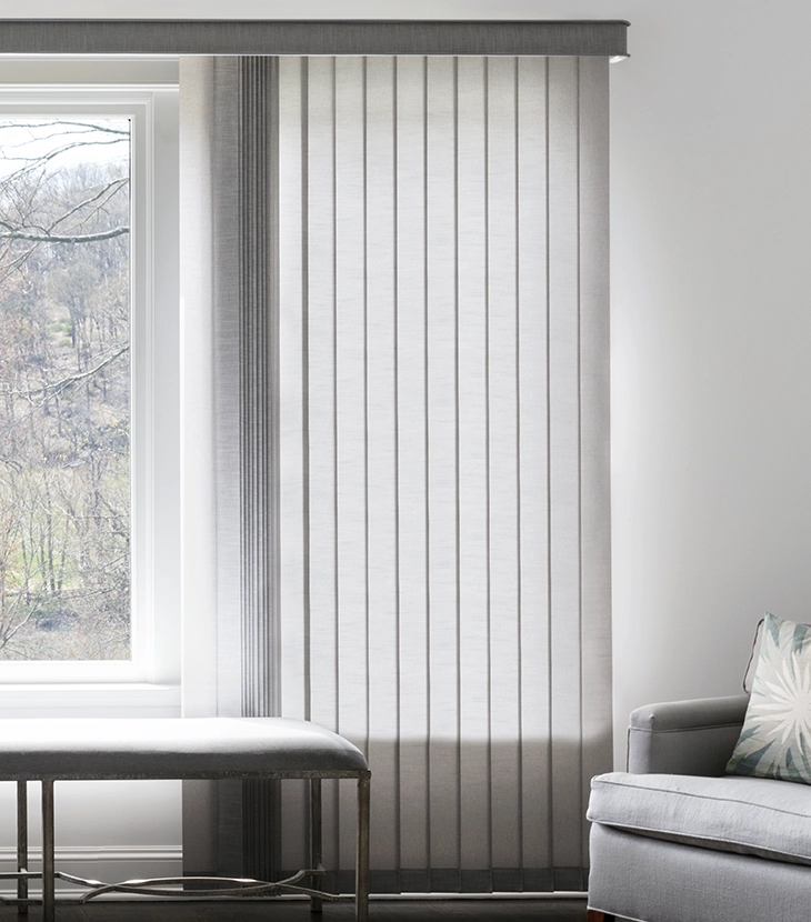 Light filters through light grey fabric vertical blinds in a large modern bedroom.