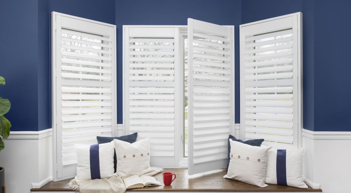 A bay window is covered with white indoor shutters with the backdrop of a dark blue wall.