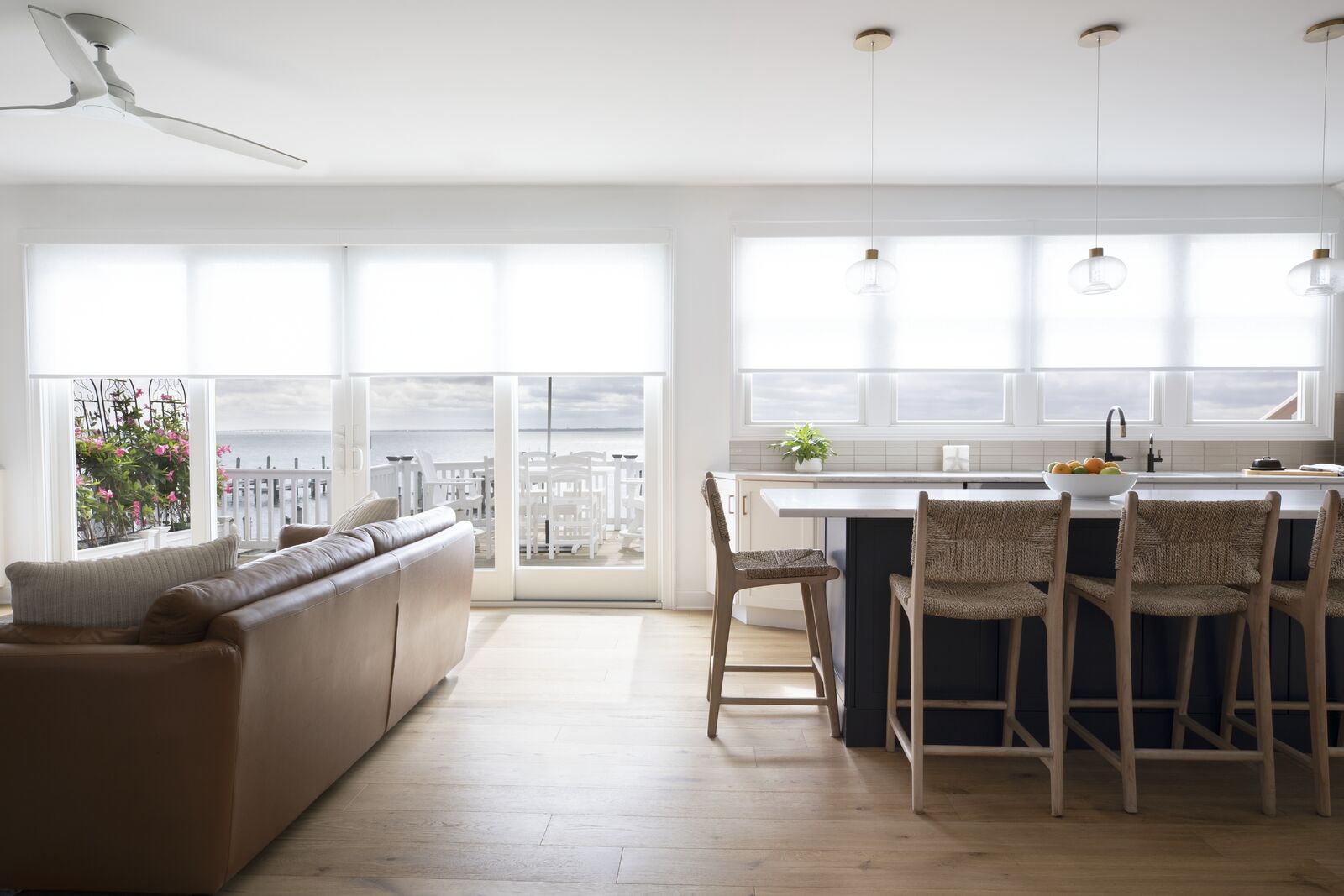 Bright and airy open-concept living room and kitchen with solar shades on large windows and sliding doors, featuring a tan leather sofa, woven bar stools, and a modern white island overlooking a waterfront view. Bright and airy open-concept living room and kitchen with solar shades on large windows and sliding doors, featuring a tan leather sofa, woven bar stools, and a modern white island overlooking a waterfront view.