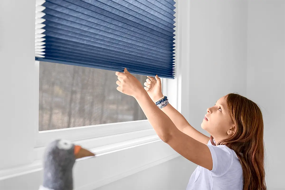 A young girl reaches up to pull down blue cellular shades A young girl reaches up to pull down blue cellular shades