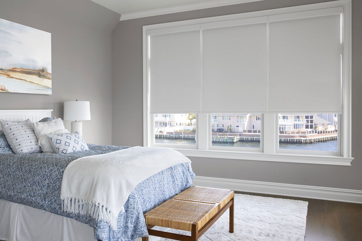 Blackout white roller shades cover three side-by-side windows in a modern waterfront bedroom Blackout white roller shades cover three side-by-side windows in a modern waterfront bedroom